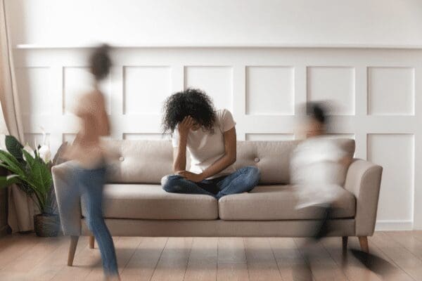 woman sitting on couch with head in hands, as children run around her
