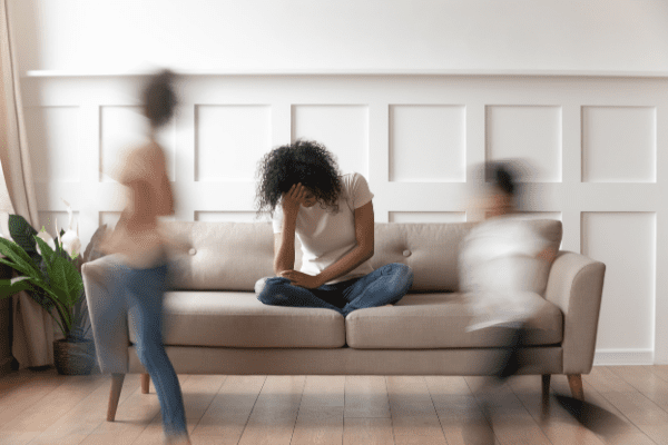 woman sitting on couch with head in hands, as children run around her